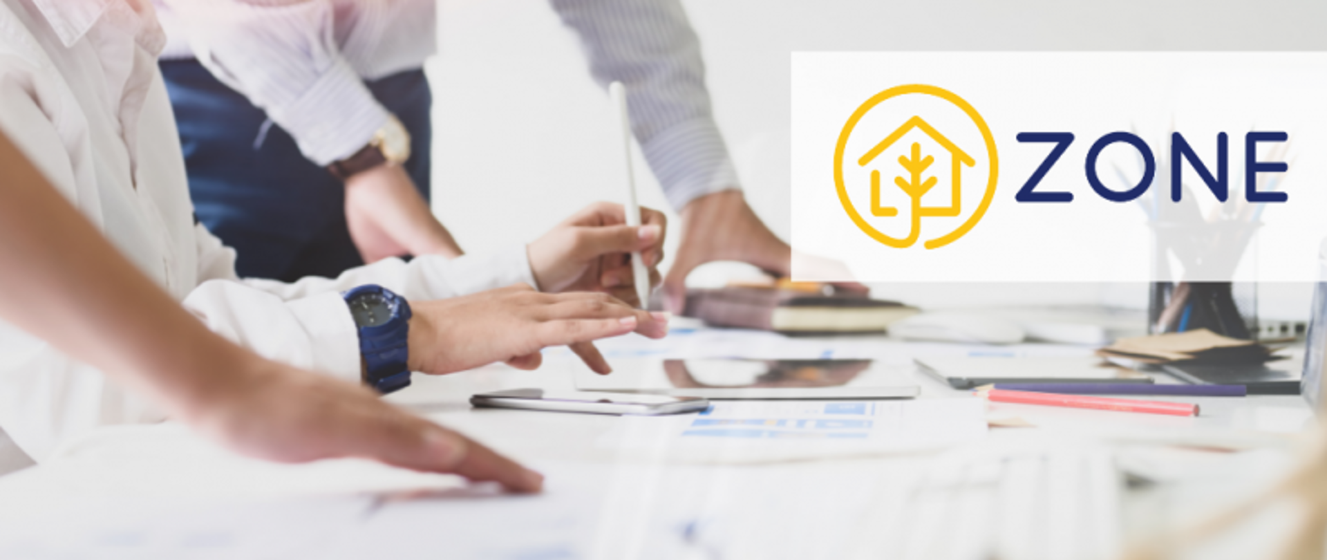Grupa osób pracująca przy stole z dokumentami i urządzeniami elektronicznymi; jedna osoba wskazuje coś długopisem, po prawej stronie widoczne logo „ZONE” z ikoną domu i drzewa.
