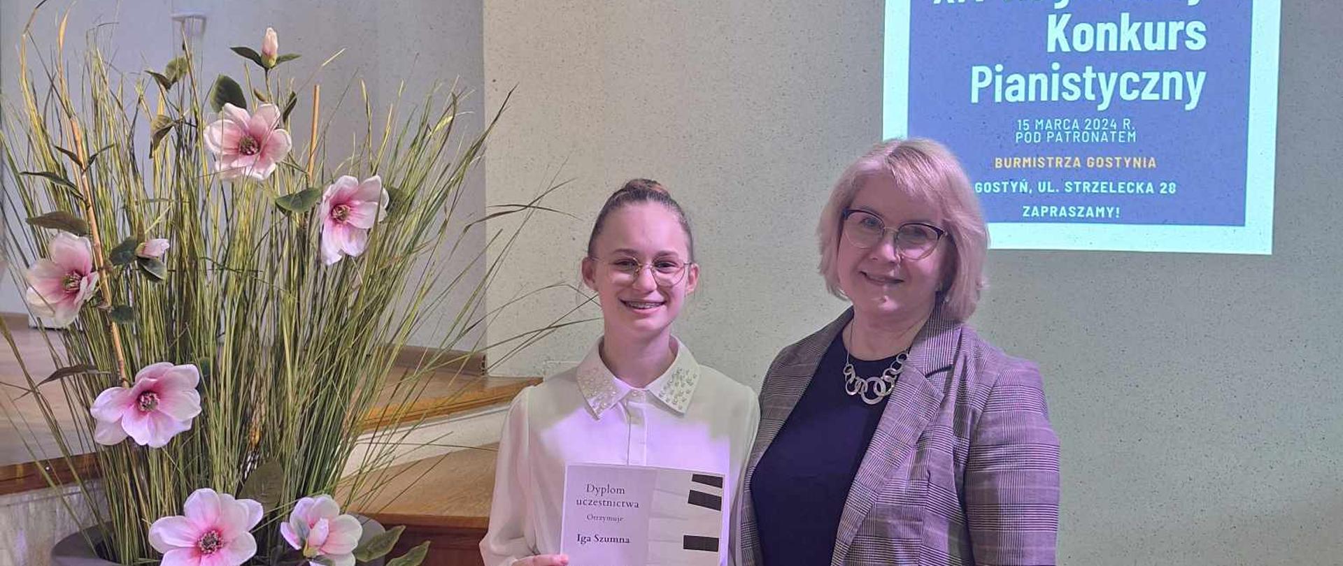 Uczennic Iga Szumna stoi wraz ze swoją nauczycielką Małgorzatą Mudry-Malajka. Dziewczynka trzyma dyplom. Z boku duża donic z kwiatami. W tle napis - XIV Regionalny Konkurs Pianistyczny.