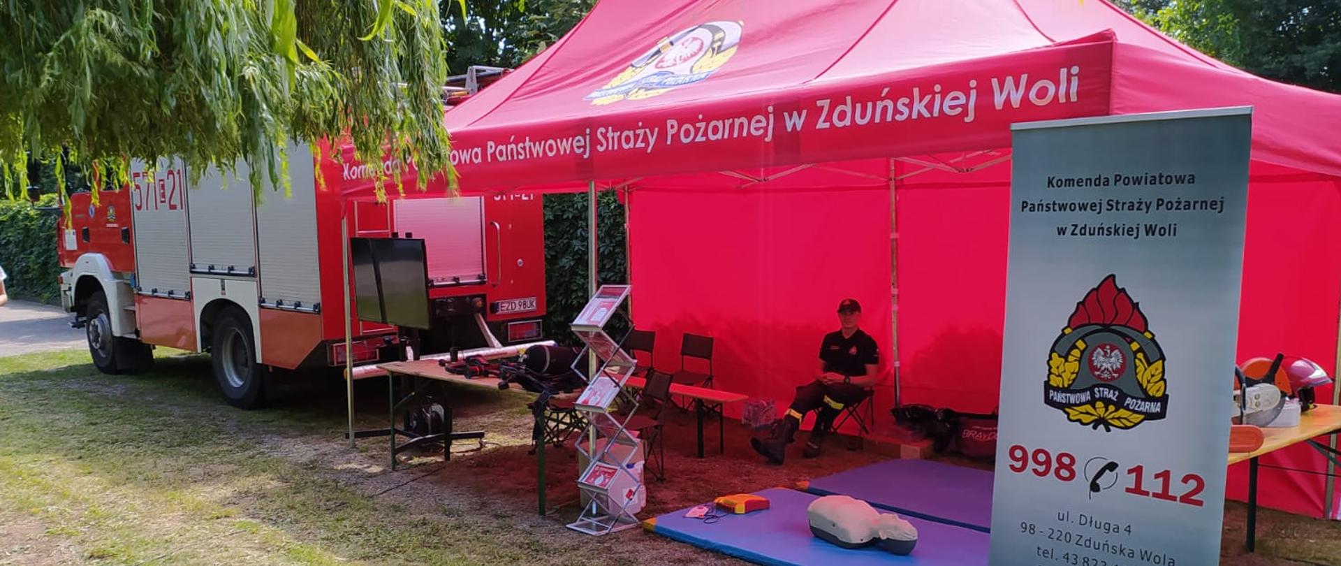 Stoisko PSP pod namiotem pod którym znajdują się sprzęty wykorzystywane przez strażaków podczas działań oraz fantom do szkolenie z RKO. W tle widoczny duży samochód pożarniczy. 