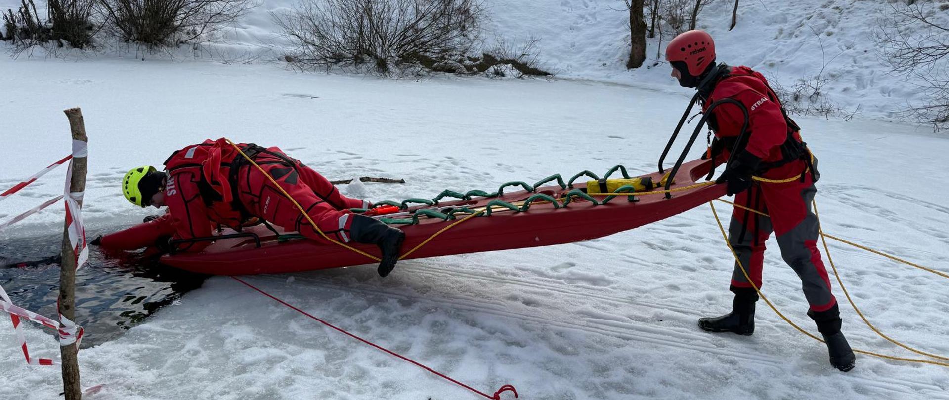 Zdjęcie przedstawia trzech strażaków w skafandrach suchych. Jeden ze strażaków stoi na lodzie i trzyma sanie z tyłu. Drugi na nich leży i podejmuje poszkodowanego z wody którym jest również strażak . 