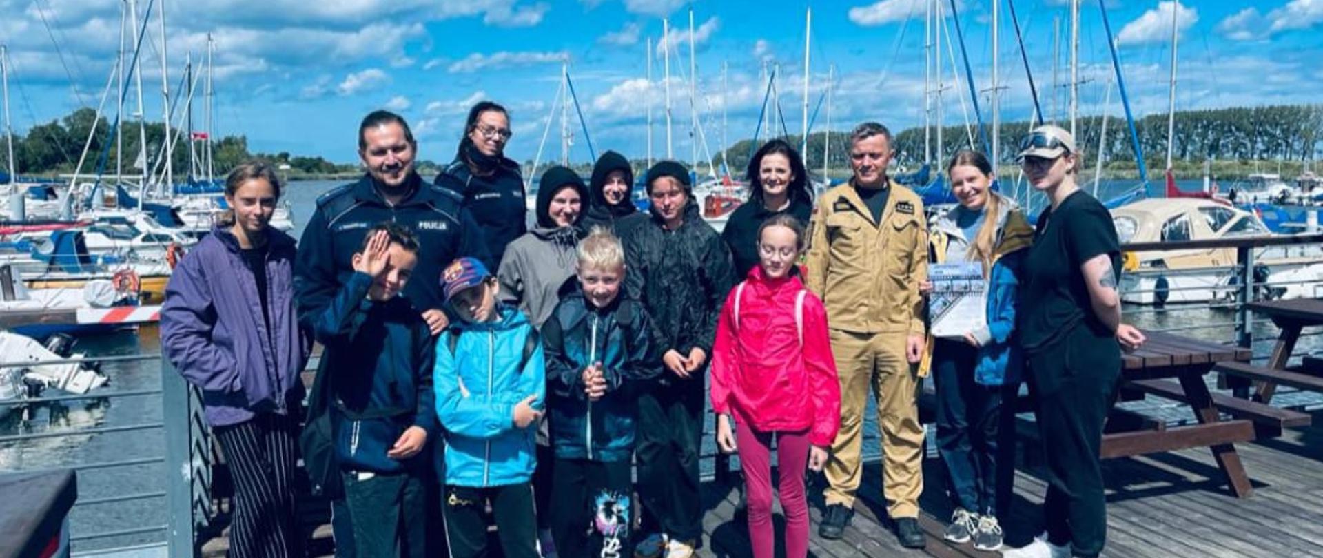 Zdjęcie przedstawia strażaków i policjantów z grupą osób wypoczywających nad wodą