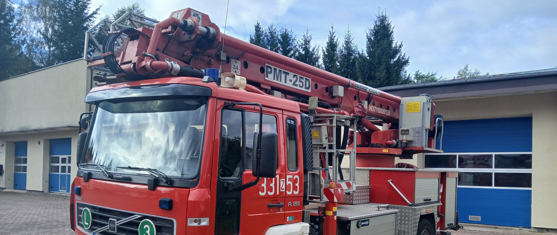 Czerwony samochód pożarniczy typu podnośnik hydrauliczny SH 25 marki Volvo stoi przed budynkiem jednostki straży pożarnej. Na pojeździe widoczny jest numer operacyjny 331Z53 oraz oznaczenia jednostki PSP. W tle widać garaże z niebieskimi bramami i drzewa.