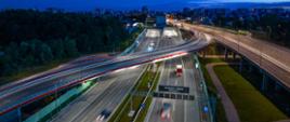 Nocny widok z lotu ptaka na wielopasmową trasę ekspresową z estakadą i wlotem do tunelu; na jezdniach widać świetlne smugi przejeżdżających samochodów, a w tle rozciąga się panorama miasta.