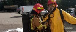 Strażacy w trakcie ćwiczeń scenariuszy chemicznych. Jeden strażak w ubraniu specjalnym, drugi w ubraniu przeciwochlapaniowym. 
