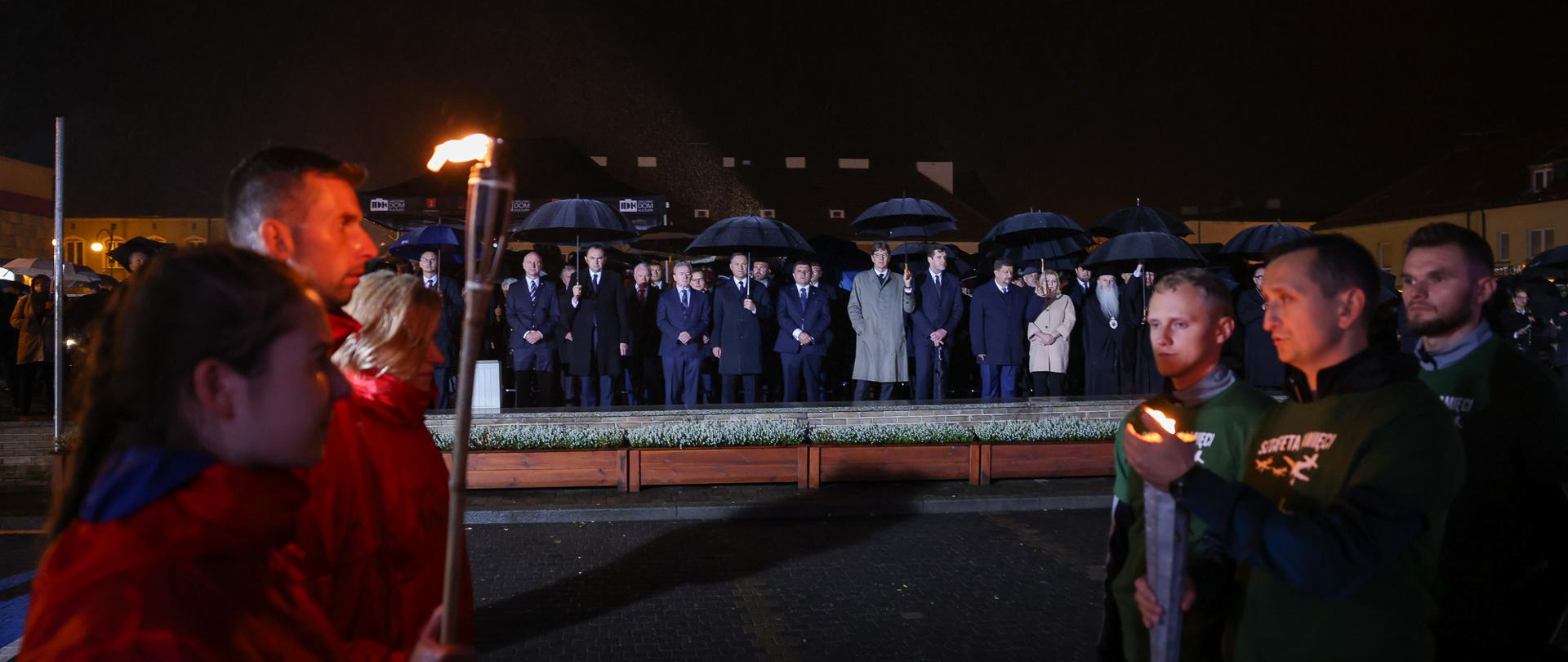 Na pierwszym planie grupa młodych osób. Po lewej stronie ubranych w czerwone kurtki. Jedna osoba trzyma w dłoni pochodnię. PO prawej stronie młodzież ubrana w zielone koszulki. Jedna osoba trzyma w ręku znicz. W tle w szeregu stoi grupa osób ubrana w płaszcze i garnitury. Nad głowami trzymają parasolki. W centrum prezydent Andrzej Duda. 