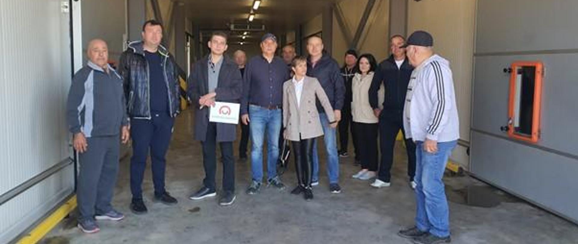 Kilkanaście osób pozuje do zdjęcia w magazynie lub chłodni. Jeden z nich trzyma znak z logo Polskiej pomocy.