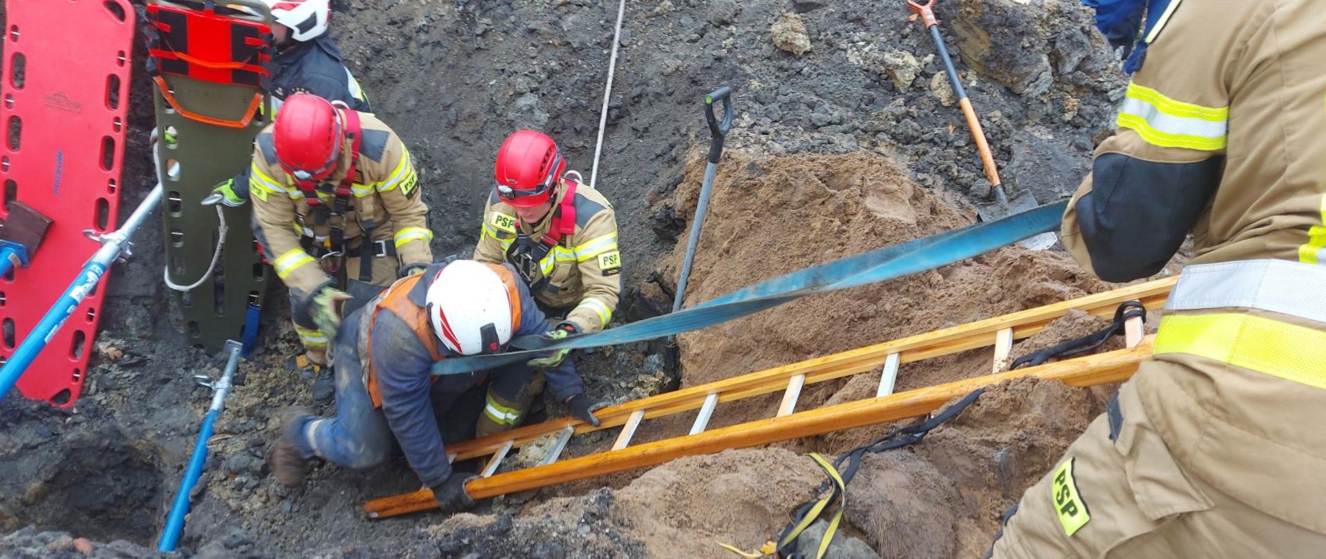 Na zdjęciu widać mężczyznę, który wychodzi po drabinie z wykopu. Strażacy asekurują poszkodowanego. 