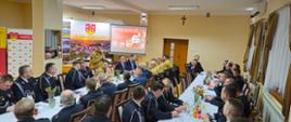 zdjęcie przedstawia strażaków w ubraniach wyjściowych oraz przemawiającego Komendanta, w tle projektor