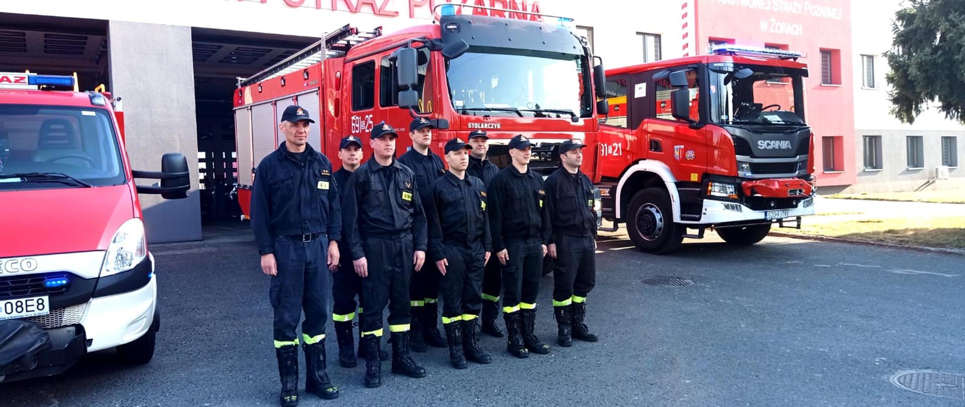 Strażacy stojący w dwuszeregu na tle samochodów trzech ustawionych przodem strażackich.
Za strażakami i samochodami budynek strażnicy.