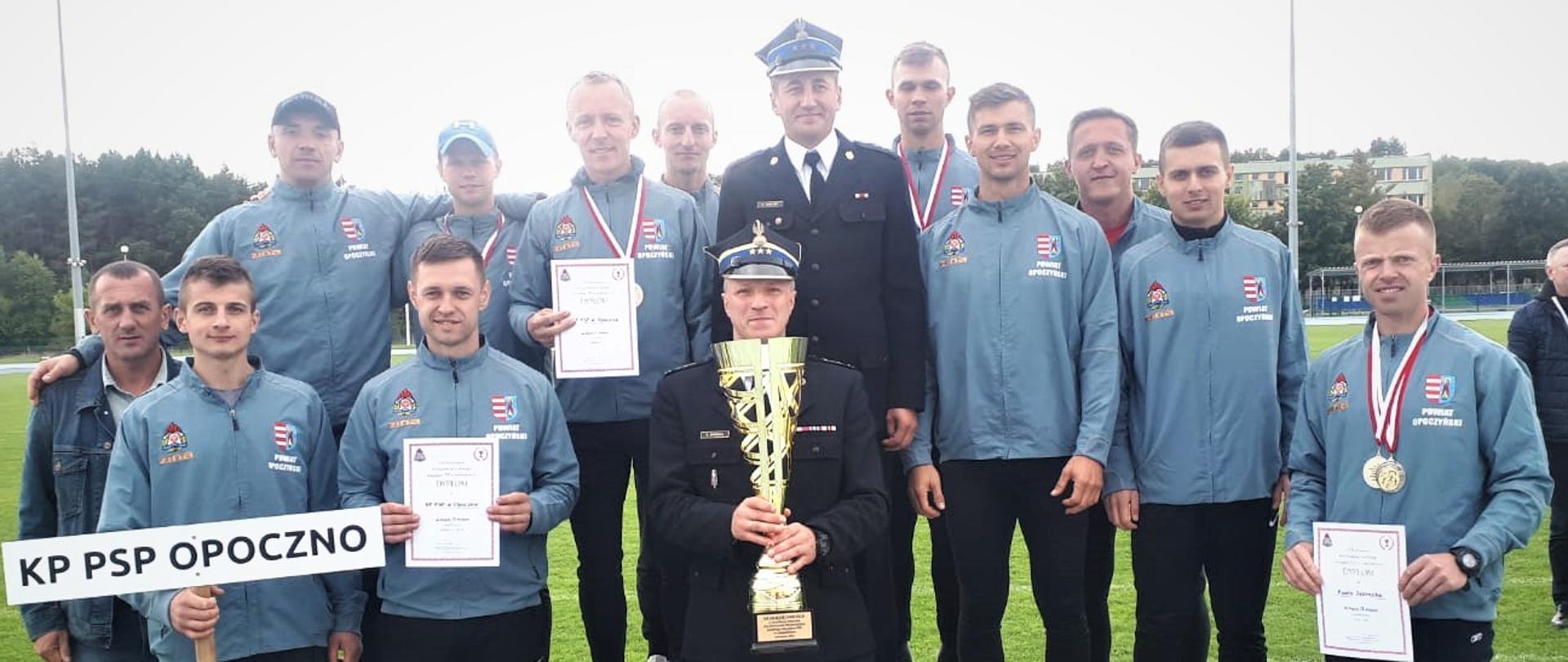 Drużyna strażaków z Komendy Powiatowej PSP w Opocznie w szarych dresach reprezentacyjnych. 10 strażaków stoi w górnym rzędzie, 4 strażaków stoi w dolnym rzędzie. W środku stoi Komendant Powiatowy PSP w Opocznie i trzyma zdobyty puchar. Pozostali strażacy trzymają dyplomy i tablice z napisem KP PSP Opoczno. W tle boisko sportowe. 