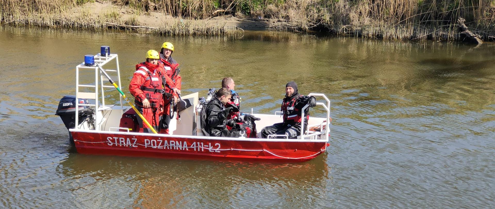 Na wodzie stoi czerwona lódź strażacka a w ładzi pięciu strażaków. Dwóch w czerwonych kombinezonach i żółtych hełmach i trzech w czarnych skafandrach do nurkowania