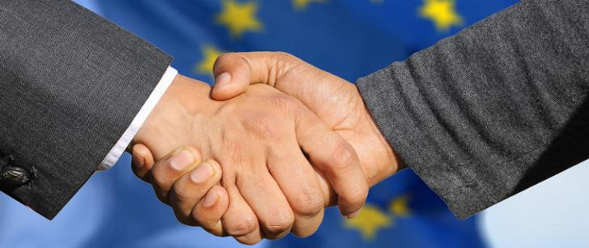 Dwie uściśnięte męskie dłonie na tle unijnej flagi.
