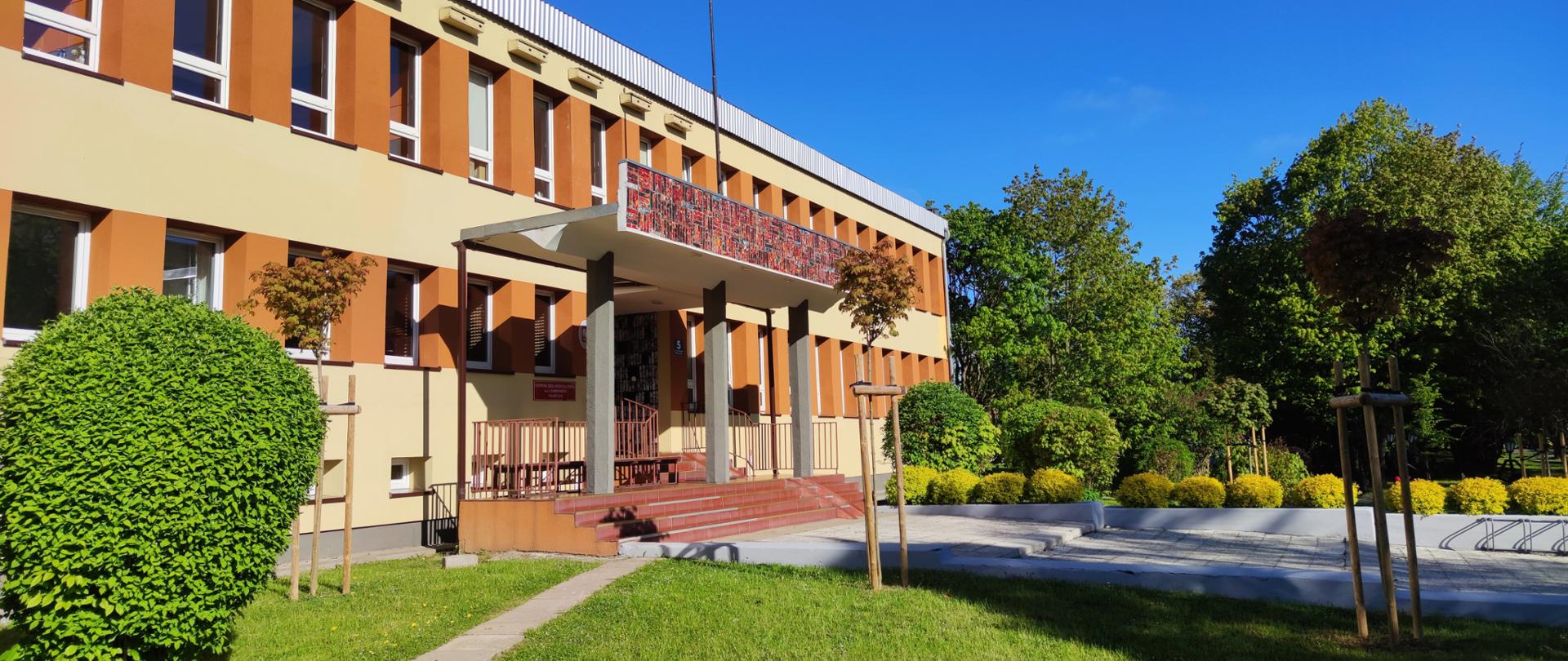 na zdjęciu widoczny jest budynek szkoły na tle niebieskiego nieba, otoczenie zielone drzewa i krzewy