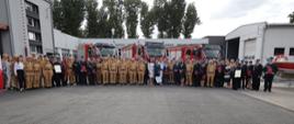 Wspólne zdjęcie funkcjonariuszy PSP, zaproszonych gości wraz z przedstawicielami jednostek OSP. W tle widoczne są przekazane samochody ratowniczo-gaśnicze oraz budynki Komendy Wojewódzkiej PSP w Opolu.
