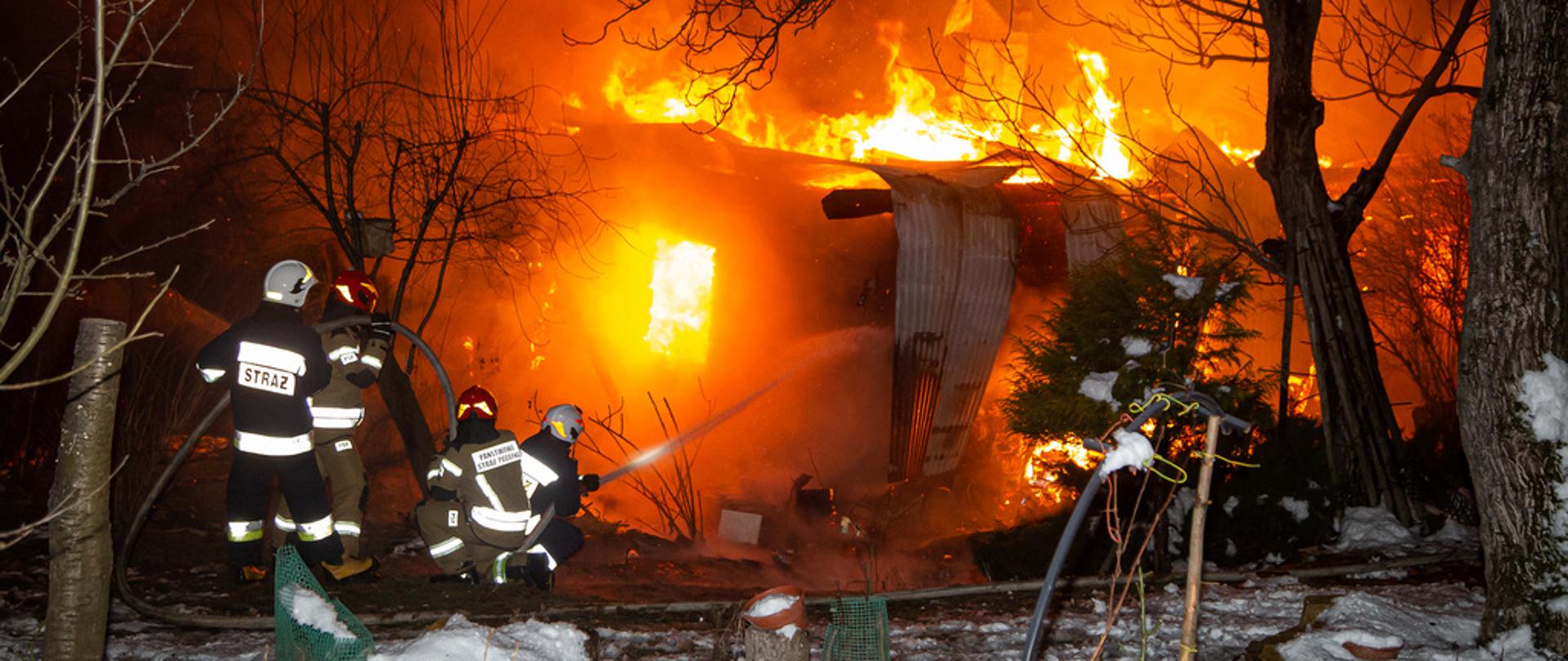 Na zdjęciu w porze nocnej widoczni czterej strażacy gaszący pożar budynku mieszkalnego. Warunki zimowe, śnieg, strażacy podają prąd wody w natarciu na palący się dom.