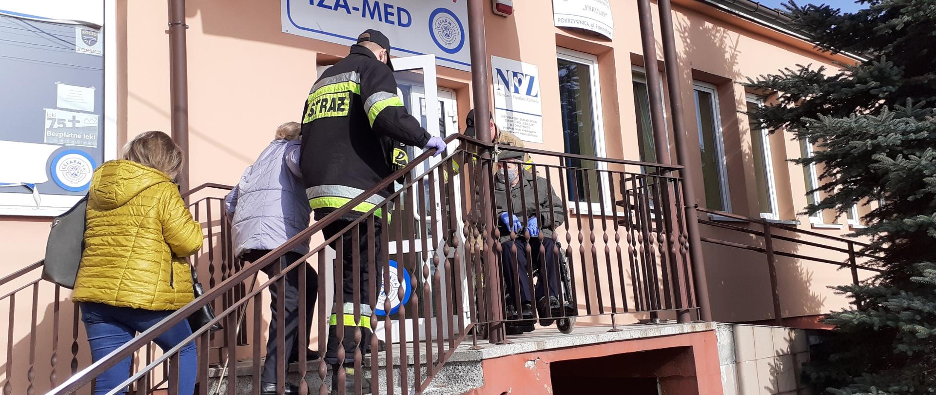 Na zdjęciu strażak pomaga wejść po schodach seniorom, którzy udają się do punktu szczepień przeciwko COVID-19