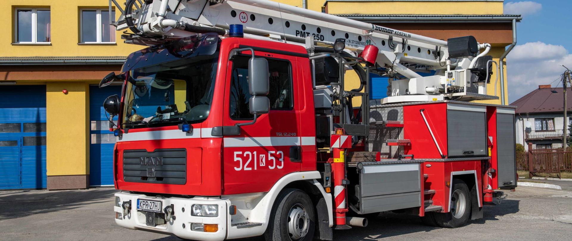 Specjalny samochód z hydraulicznie podnoszonym koszem i wysuwaną drabiną marki MAN na tle bram wjazdowych do garażu od strony placu wewnętrznego. Podnośnik z koszem i drabina są w stanie złożonym na nadwoziu pojazdu.