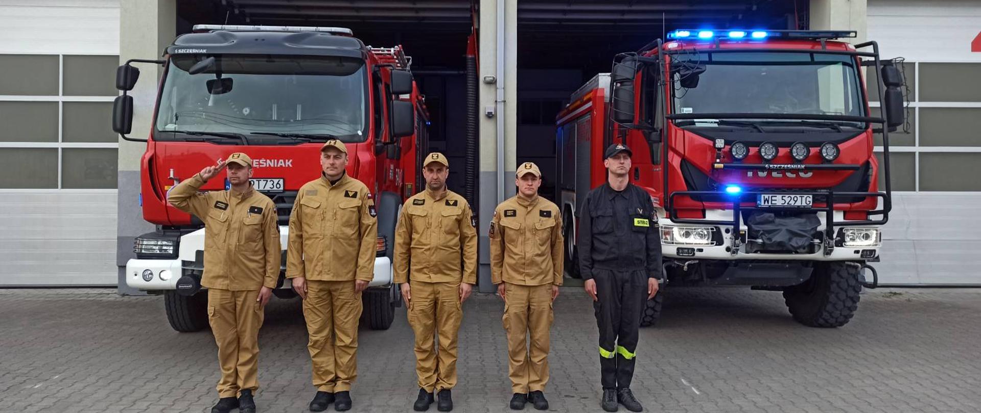 Zdjęcie przedstawia funkcjonariuszy PSP w ubraniach koszarowych koloru piaskowego i czarnego. Stojących w jednym szeregu przed czerwonym samochodami gaśniczymi. Zdjęcie wykonane przed garażem JRG.