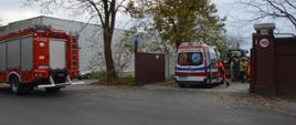 Wypadek podczas prac polowych. Teren jednego z zakładów pracy na terenie Rawicza. Widok z ulicy. Strażacy i ratownicy medycznie udzielają pomocy kobiecie z urazem ręki. Na ulicy z lewej strony widoczny jest tył wozu strażackiego, natomiast w bramie wjazdowej do zakładu stoi karetka pogotowia ratunkowego.