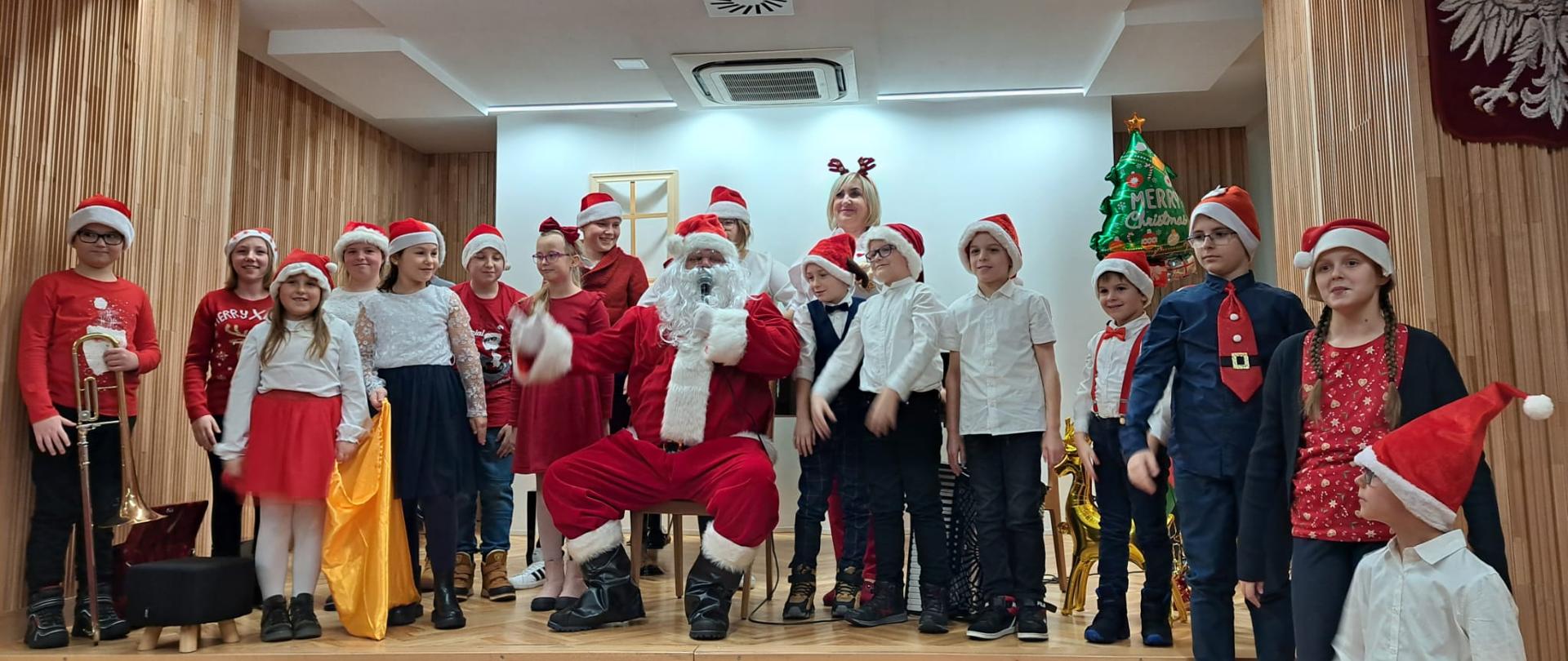 Zdjęcie grupowe przedstawia stojących na scenie wykonawców koncertu, pośrodku siedzi Mikołaj