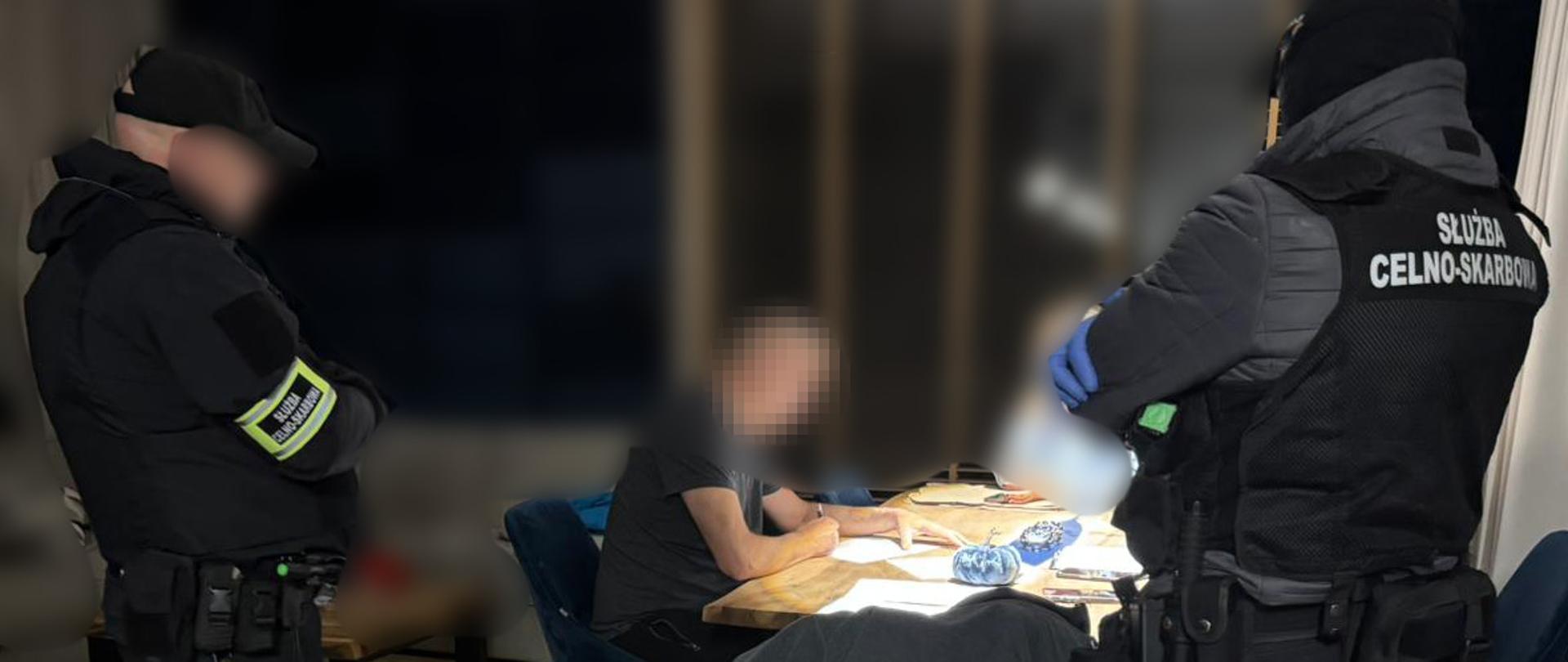 Dwóch funkcjonariuszy Służby Celno-Skarbowej stoi przy stole, przy którym siedzi osoba z zamazanym wizerunkiem.