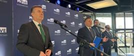 Zdjęcie z boku, na tle granatowej ścianki z napisem MOVE.stoi minister Wieczorek, obok niego trzech mężczyzn w garniturach.