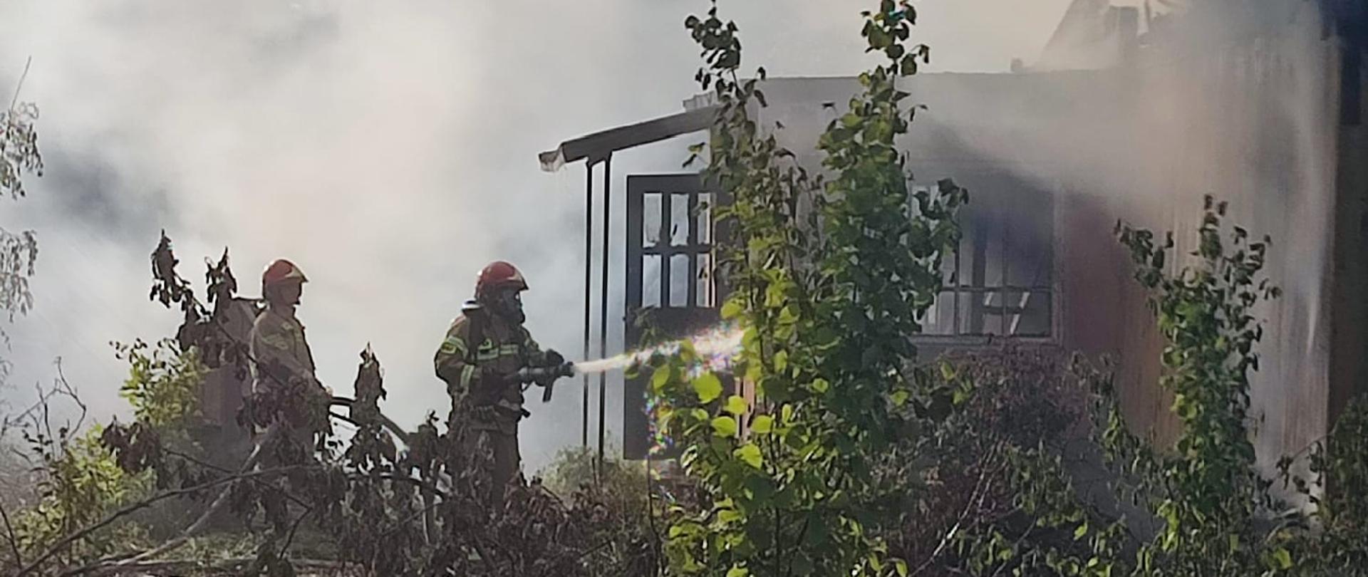 Zdjęcie przedstawia przód opuszczonego budynku mieszkalnego osnutego dymem i parą wodną, po lewej rota strażaków podająca wodę do wnętrza domu przez drzwi wejściowe.