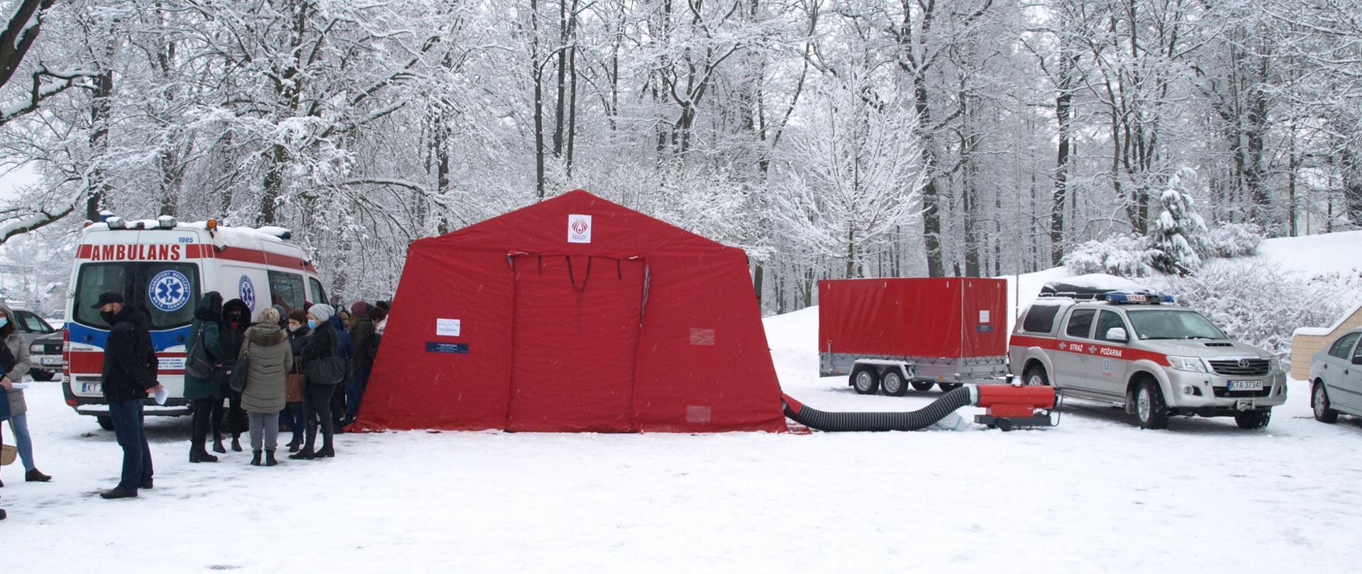 Na zdjęciu widać park w miejscowości Gromnik w zimowej aurze z zaśnieżonym placem przed amfiteatrem letnim oraz drzewami parkowymi. Na tle parku w centralnej części zdjęcia widać czerwony namiot stelażowy z podłączoną nagrzewnicą ciepłego powietrza. Po prawej stronie namiotu stoi srebrny pojazd pożarniczy Toyota Hilux z przyczepką do przewozu namiotu - w dyspozycji OSP Wojnicz. Po lewej stronie namiotu w kolejce stoją osoby, które zostaną poddane badaniu medycznemu w zakresie COVID19. Obok osób oczekujących na badanie (po lewej stronie) zaparkowany jest pojazd - karetka pogotowia.