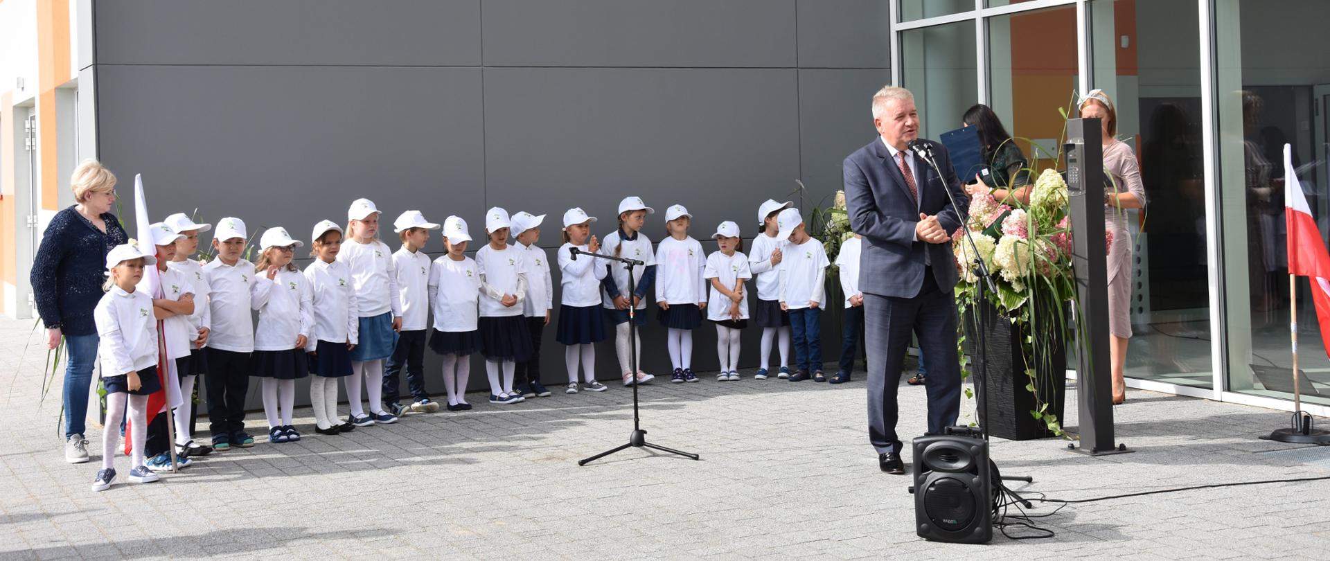 Na pierwszym planie zdjęcia, przed mikrofonem stoi ubrany w ciemny garnitur, białą koszulę i czerwony krawat mężczyzna. W tle, w szeregu stoją przedszkolaki, a tuż za nimi fragment elewacji budynku, w którym znajduje się żłobek i przedszkole. 
