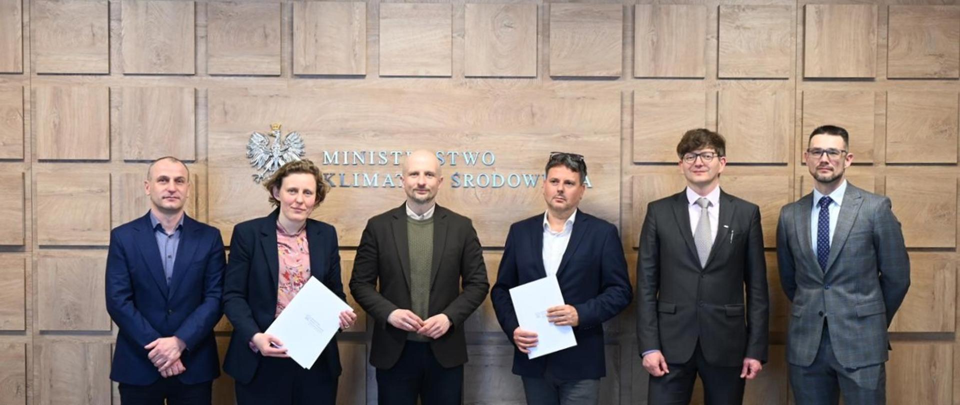 Grupa przedstawicieli Ministerstwa Klimatu i Środowiska oraz Centralnego Azylu dla Zwierząt stoi przed ścianą z godłem RP i logotypem ministerstwa, prezentując podpisane dokumenty.
