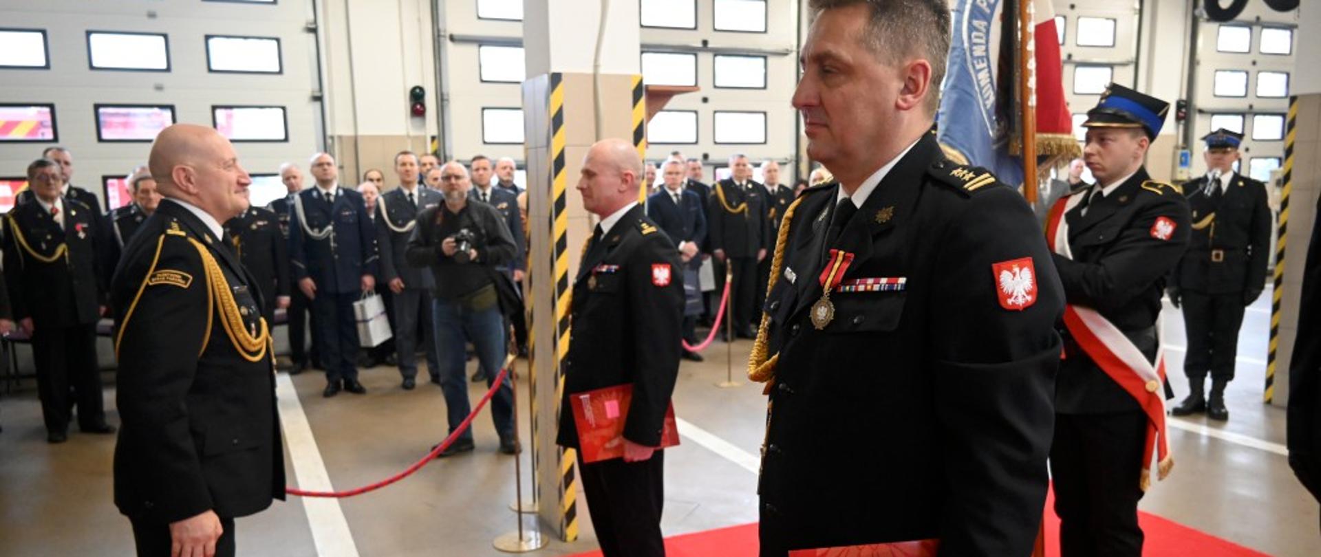 Na czerwonym dywanie stoją dwaj strażacy w mundurze galowym. Przed nimi trzeci, przyjmuje meldunek. Za nimi widać strażaka ze sztandarem. W tle kilkadziesiąt osób - załoga, goście.