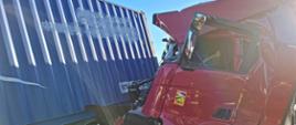 Zdjęcie przedstawia samochody ciężarowe po wypadku. Jeden pojazd z uszkodzoną kabiną koloru czerwonego uderzył w bok naczepy. 