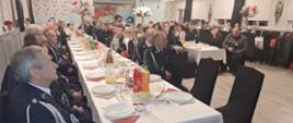 Wnętrze sali wiejskiej. W trzech rzędach ustawione są stoły, przy których po obu stronach siedzą uczestnicy spotkania. Sa to zarówno strażacy OSP i PSP w mundurach wyjściowych oraz osoby cywilne - kobiety, mężczyźni i dzieci. Na stołach znajdują sie talerze, sztućce, napoje i potrawy. Sala jestprzyozdobiona kwiatami i balonami. 