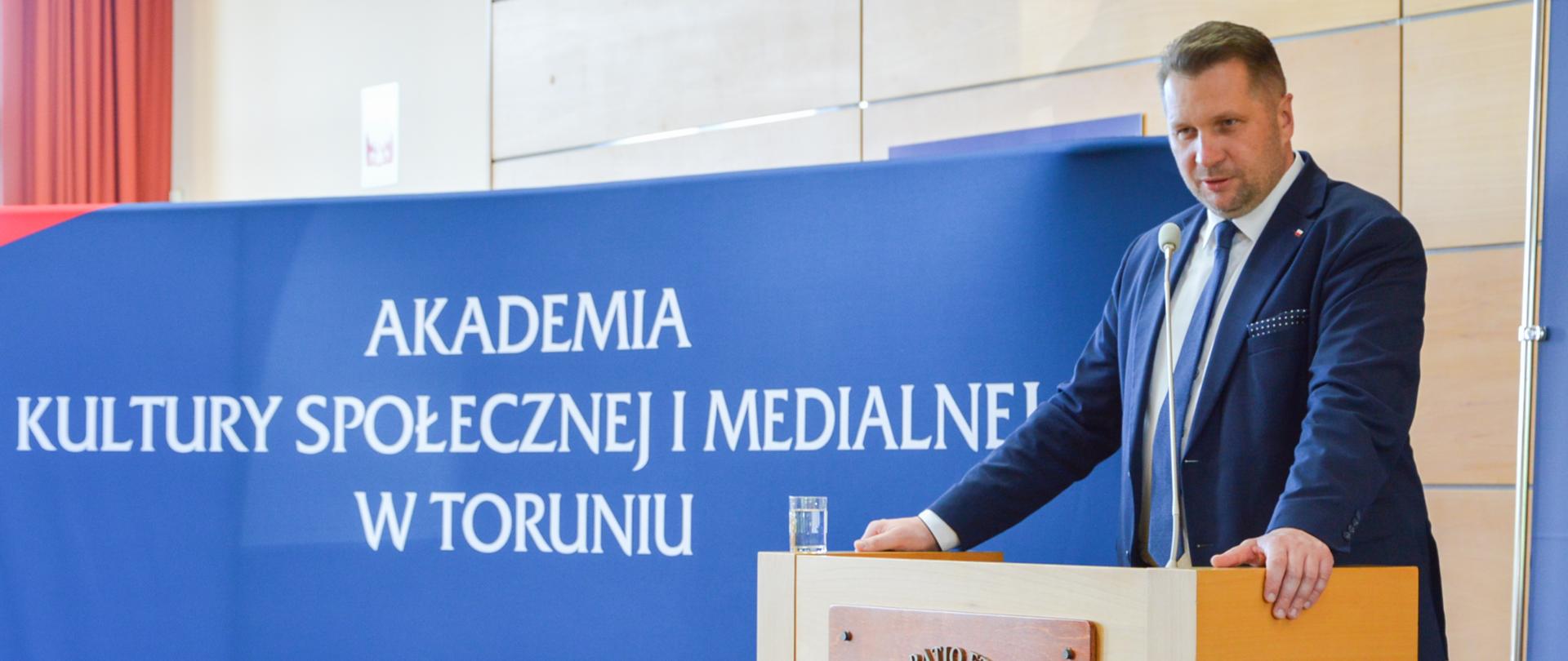Minister Przemysław Czarnek przemawia podczas wykładu. Stoi przy mównicy. Za sobą ma baner z napisem Akademia Kultury Społecznej i Medialnej w Toruniu