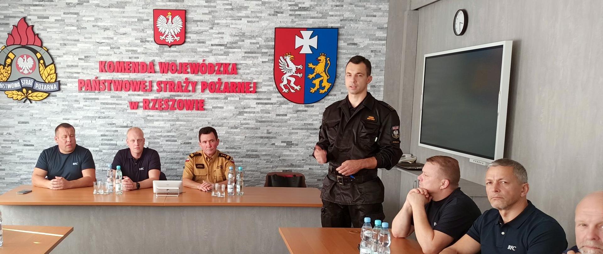 Zdjęcie zrobione w sali konferencyjnej. W sali, przy stołach konferencyjnych siedzą są strażacy. Wśród nich funkcjonariusz Państwowej Straży Pożarnej stopniu starszego brygadiera i kapitana. Strażacy uczestniczą naradzie.