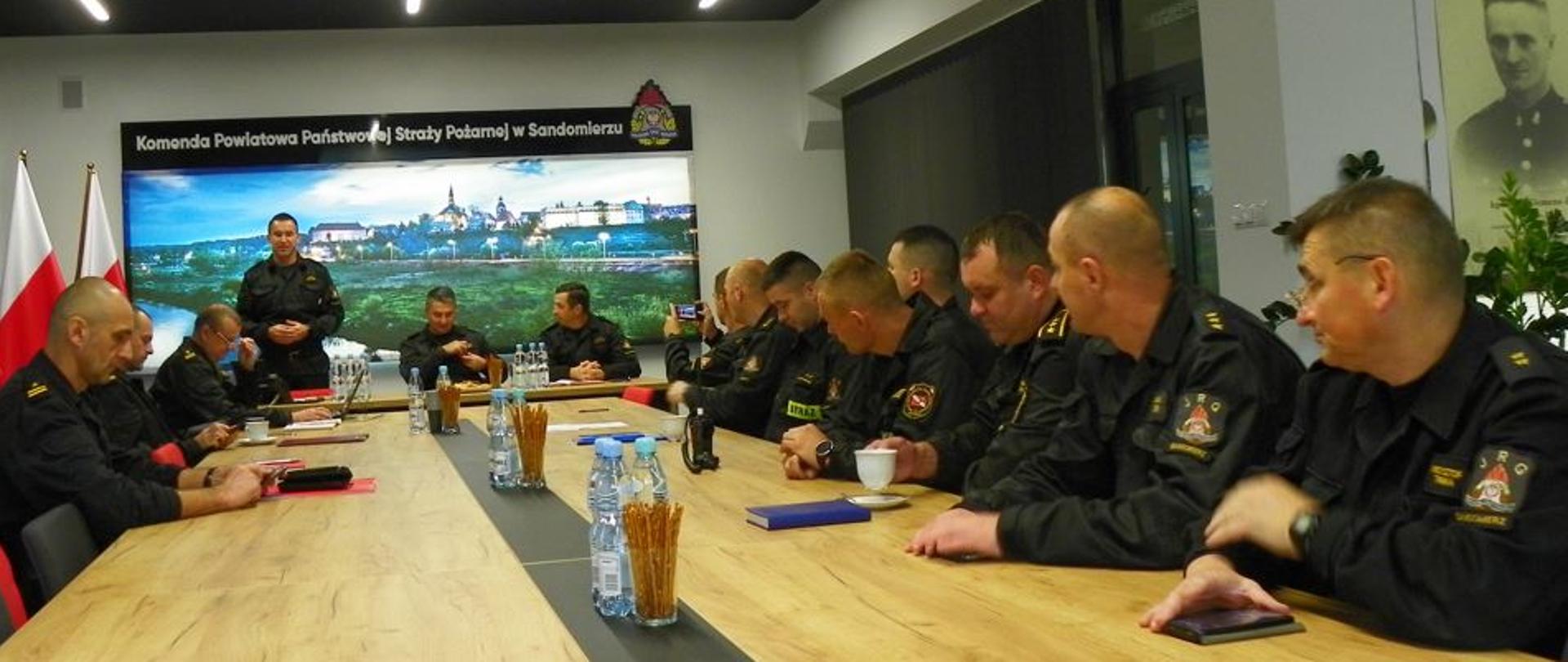 Na zdjęciu widać siedzących w pomieszczeniu narad przy stole prezydialnym grupę funkcjonariuszy PSP w umundurowaniu dowódczo-sztabowym.