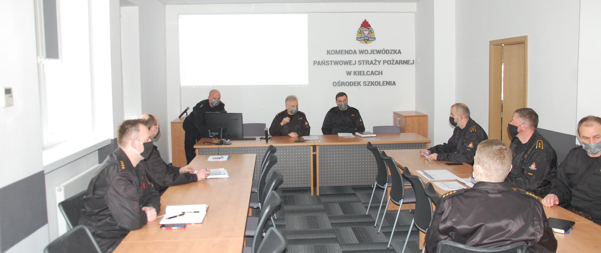 Zdjęcie dotyczy narady szkoleniowej grup specjalistycznych. W sali narad przy stołach siedzi 8 funkcjonariuszy w mundurach oraz jeden funkcjonariusz, który stoi przy komputerze i przedstawia prezentację. Na zdjęciu Zastępca Świętokrzyskiego Komendanta Wojewódzkiego Państwowej Straży Pożarnej starszy brygadier Jarosław Piotrowski, koordynatorzy poszczególnych grup specjalistycznych województwa świętokrzyskiego oraz kadra Ośrodka Szkolenia Komendy Wojewódzkiej Państwowej Straży Pożarnej w Kielcach. W tle z projektora wyświetlane tabele szkoleniowa poszczególnych grup specjalistycznych