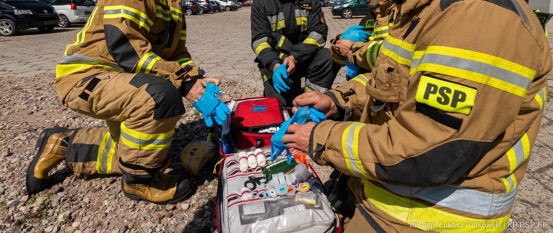 Na zdjęciu strażacy zakładający rękawiczki ochronne oraz otwarta torba medyczna ze sprzętem.