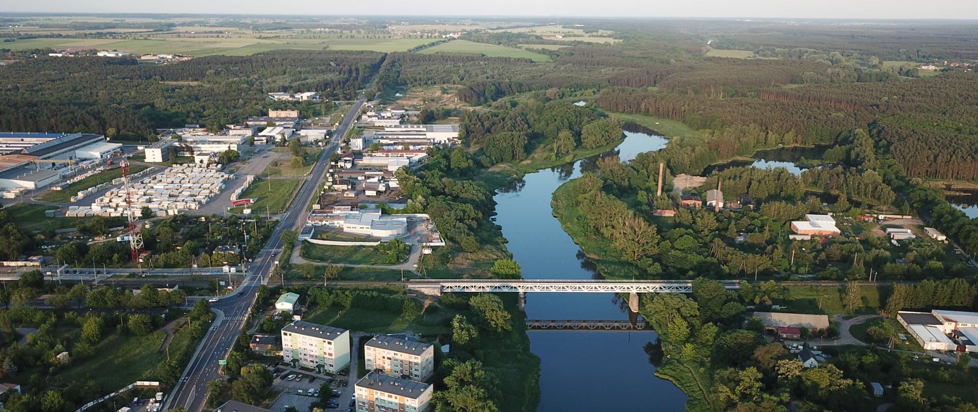 Widok z lotu ptaka na budynki mieszkalne i magazynowe, a także płynącą rzekę. Dookoła dużo zieleni: lasy, łąki. 