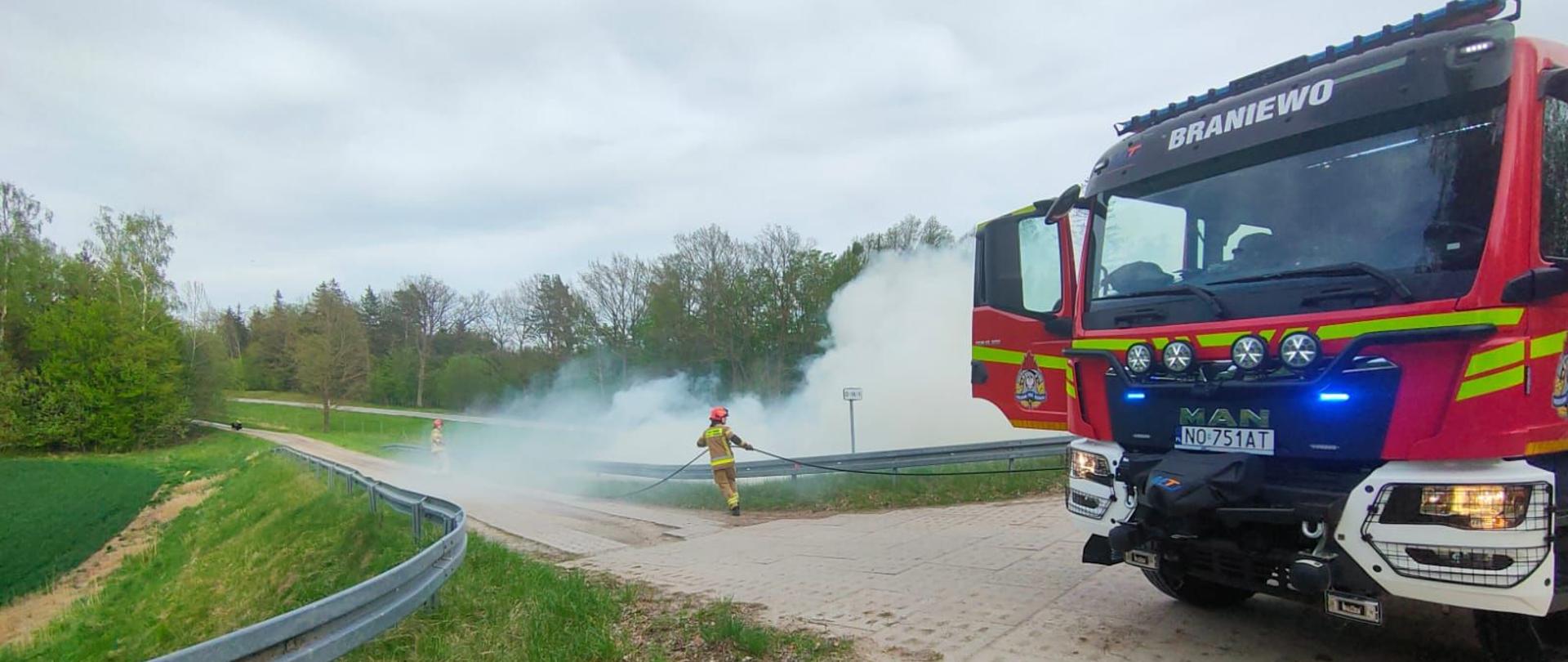 Z prawej strony przód samochodu strażackiego. Za nim dużo białego dymu. Z lewej strony tereny zielone.
