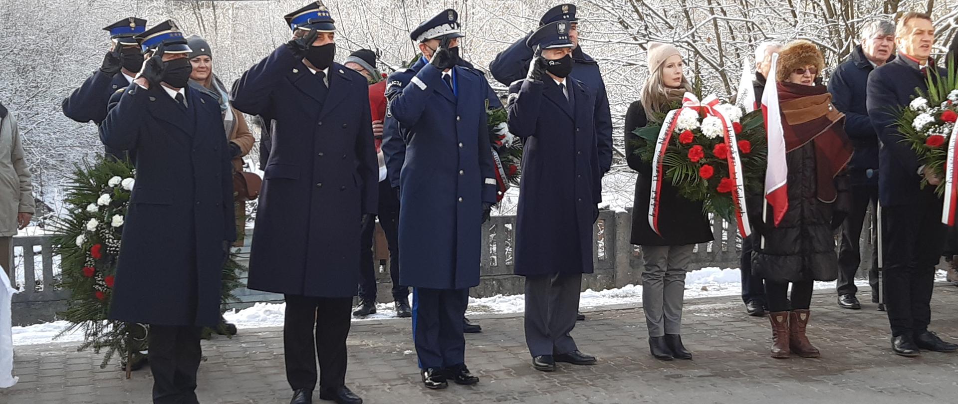 Funkcjonariusze różnych formacji mundurowych salutują w postawie na baczność. Obok nich kilka osób z bało- czerwonymi wieńcami i flagami.