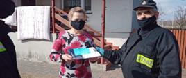 Zdjęcie przedstawia dwóch strażaków OSP, którzy wręczają kobiecie ulotkę dotyczącą szczepień przeciwko COVID-19. Zdjęcie wykonane jest na tle budynku mieszkalnego. Widać wejście do budynku oraz część ogrodzenia.