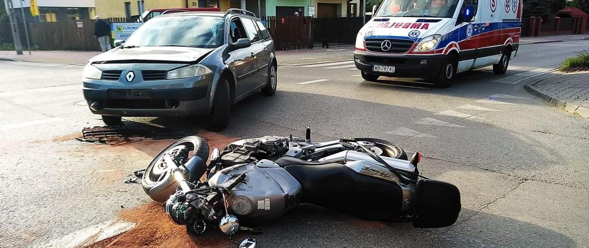 Zdjęcie przedstawia samochód osobowy marki Renault stojący na przeciwległym pasie ruchu, z uszkodzonym przodem. W odległości kilku metrów przed autem znajduje się motocykl w kolorze srebrnym leżący na boku, pojazd ma uszkodzoną przednią cześć. W oddali za tymi pojazdami uczestniczącymi w zdarzeniu znajduje się karetka pogotowia stojącą z włączoną sygnalizacją i alarmową. W głębi tych wydarzeń znajdują się dwa budynki jednorodzinnego w zabudowie bliźniaczej w kolorze żółtym oraz zieleni.