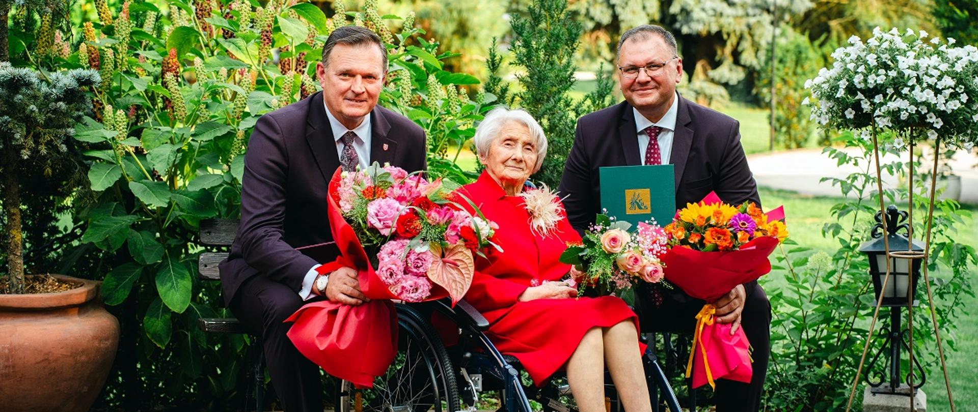 W ogrodzie siedzi starsza kobieta na wózku inwalidzkim, ubrana jest na czerwono w ręku trzyma bukiet kwiatów. Obok niej siedzą na ławce dwaj mężczyźni w garniturach, również trzymają bukiety. W tle widać bujną, zieloną roślinność.