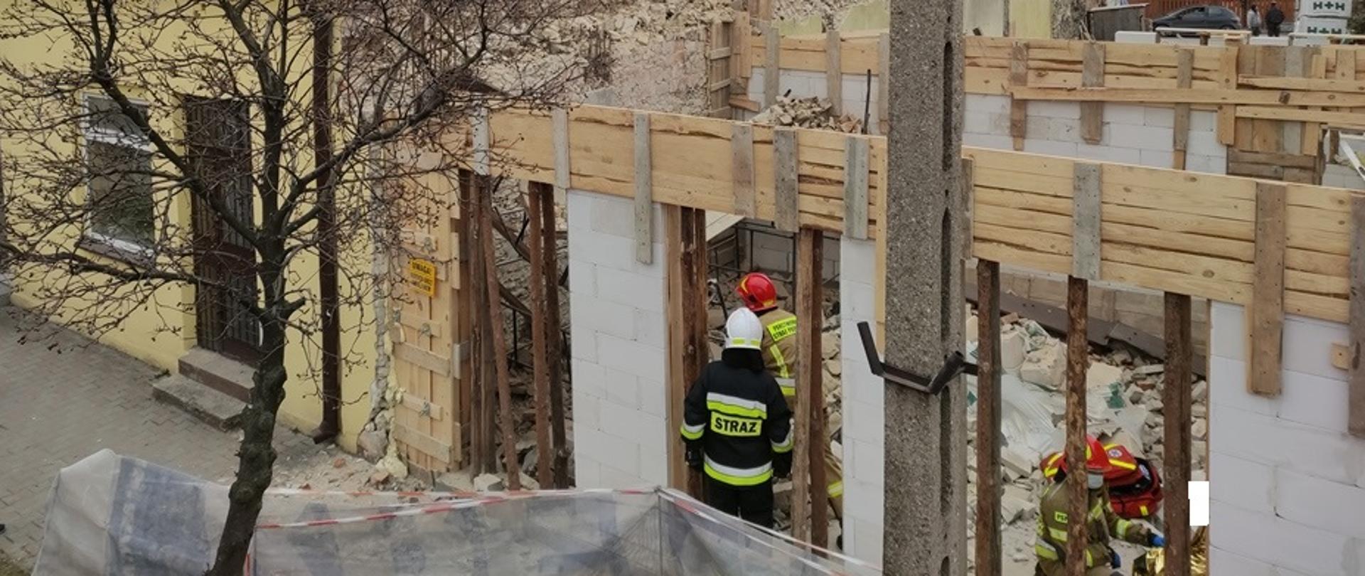 Na zdjęciu w lewej części budynek mieszkalny, uszkodzony w wyniku katastrofy budowlanej, w prawej części fotografii nowo budowany obiekt, wewnątrz strażacy prowadzący działania ratownicze, udzielanie kwalifikowanej pierwszej pomocy oraz prowadzący przerzucanie gruzowiska