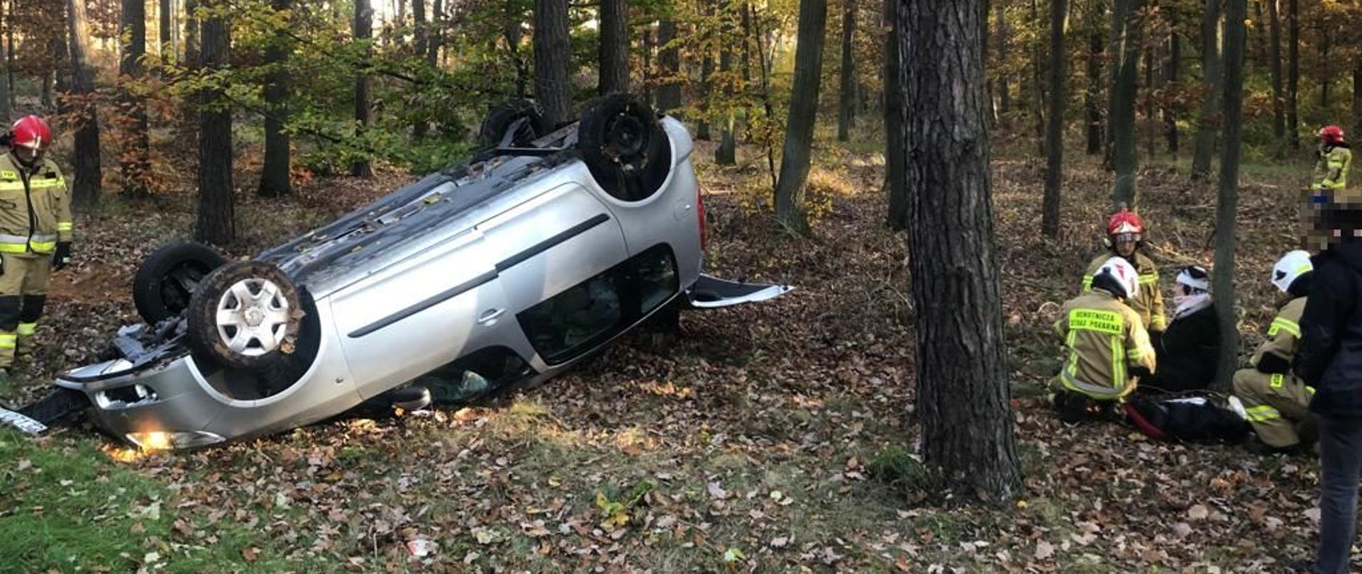 Samochód leżący na dachu, opok pojazdu strażacy udzielający pierwszej pomocy poszkodowanej kobiecie siedzącej pod drzewem.