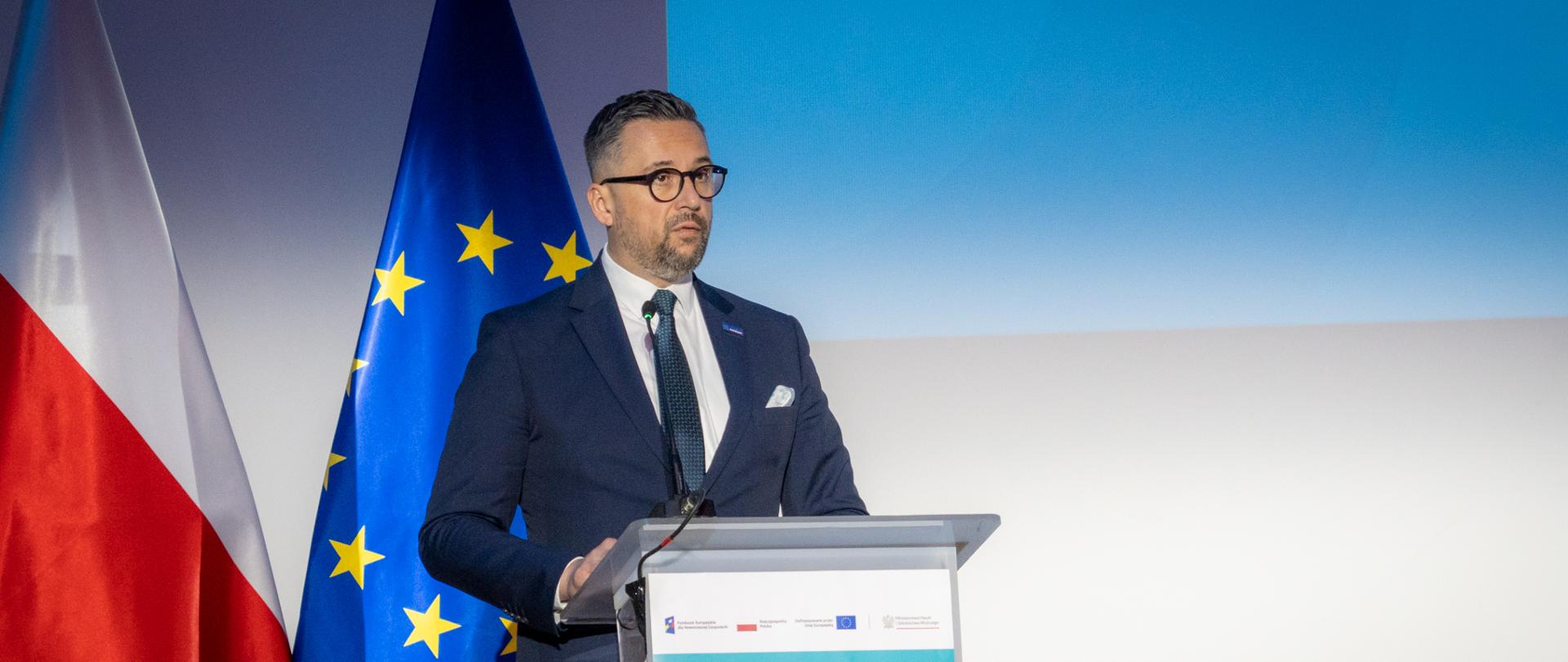Minister Kulasek stoi za mównicą i mówi do mikrofonu, za nim pod ścianą flagi Polski i UE.