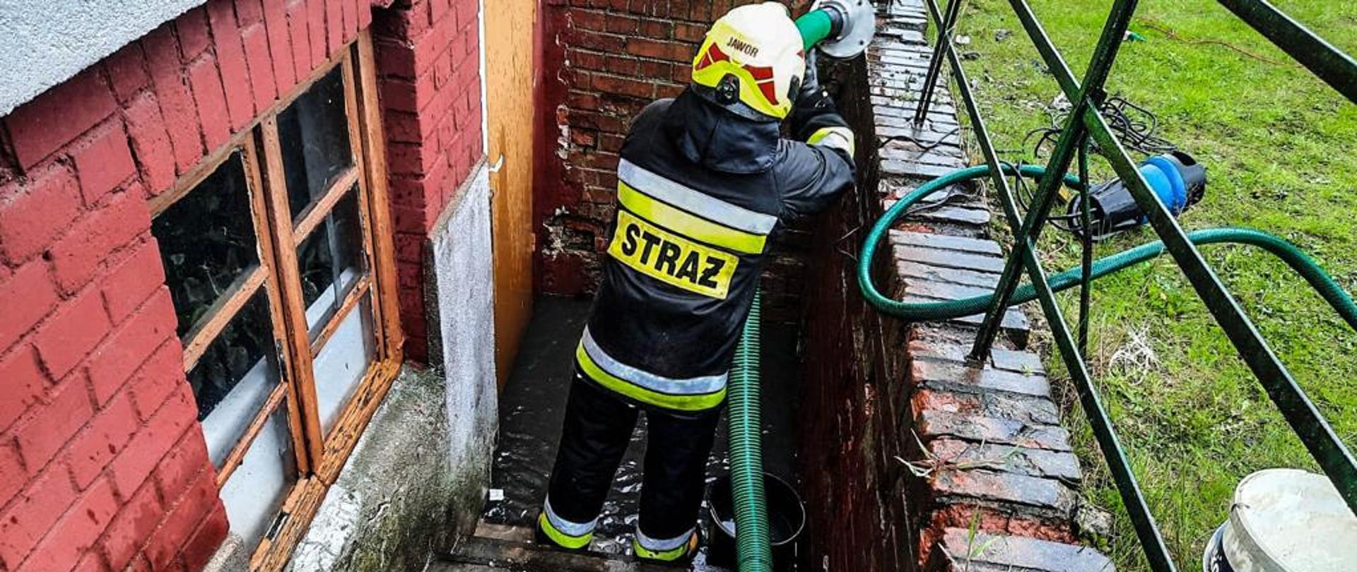 Obraz przedstawia strażaka wypompowującego wodę z zalanej piwnicy.