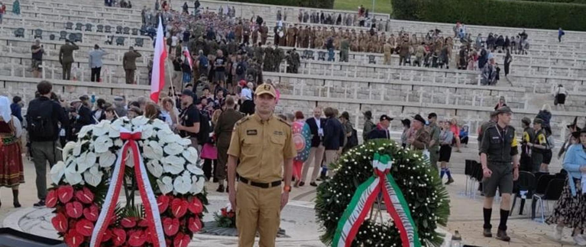 Zdjęcie pamiątkowe, mł.asp. Michał Szewczyk ubrany piaskowego koloru umundurowanie w służbowym na tle cmentarza i wielu biało-czerwonych wieńców i wiązanek. 