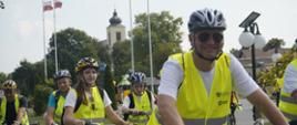 Kolumna rowerzystów w żółtych odblaskowych kamizelkach jedzie drogą, w tle wieża kościoła.